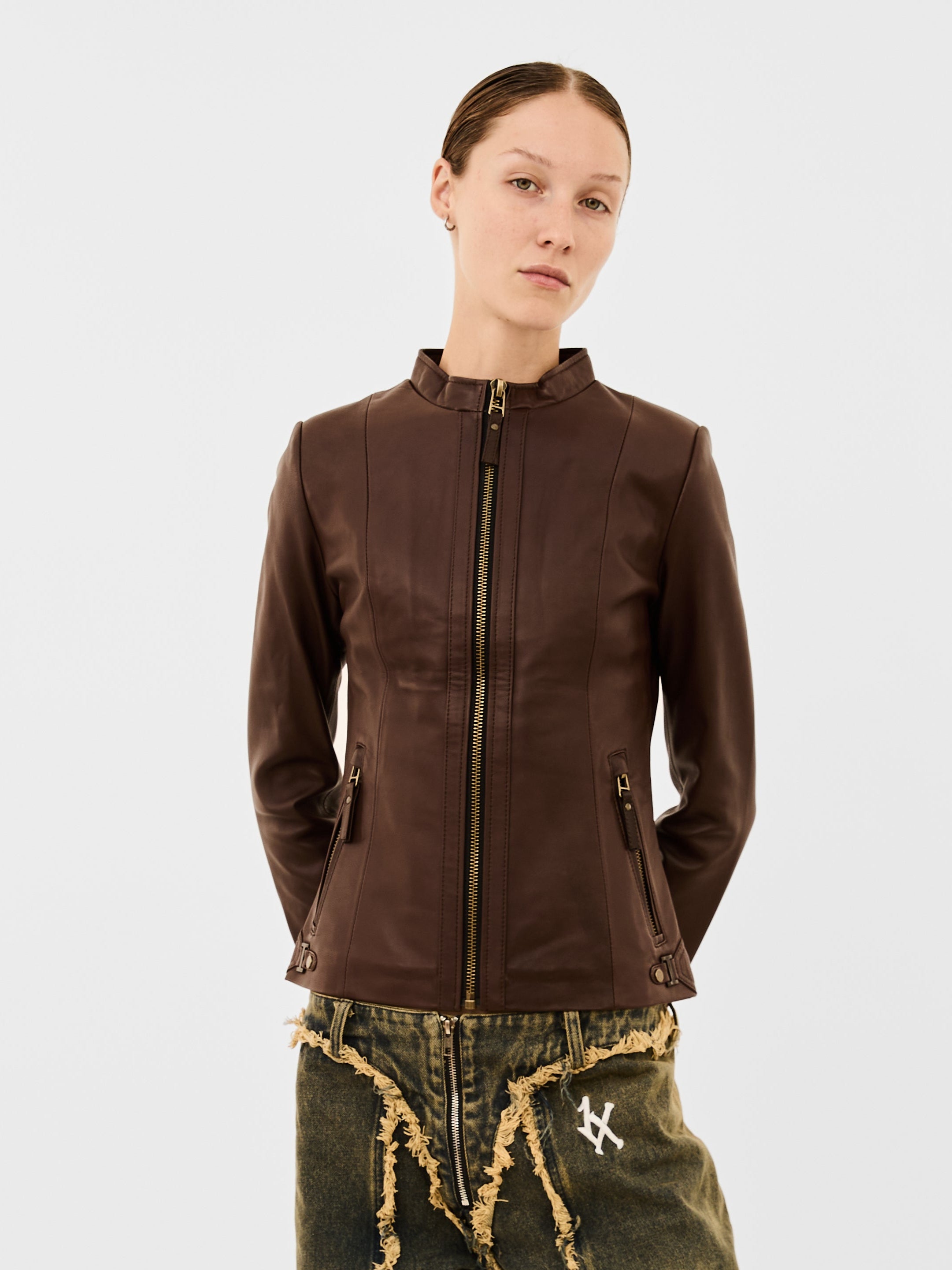 A woman wearing a brown leather jacket and distressed green jeans on a white background